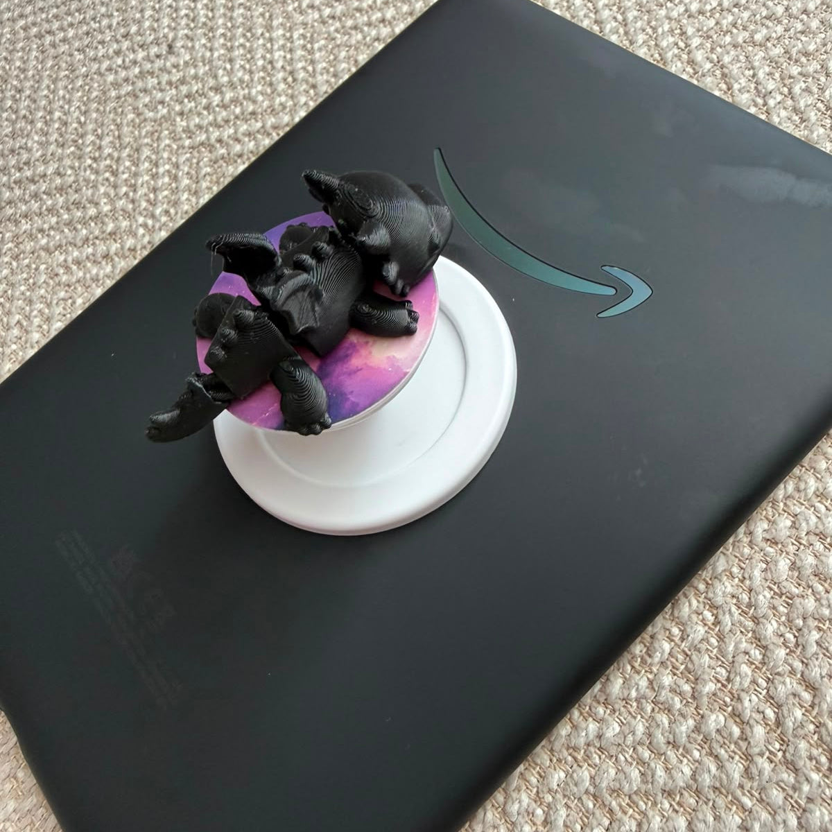 A black tablet case with a teal Amazon smile logo, resting on a beige carpet. On top of the case sits a small black figurine or decorative object placed on a round white stand.