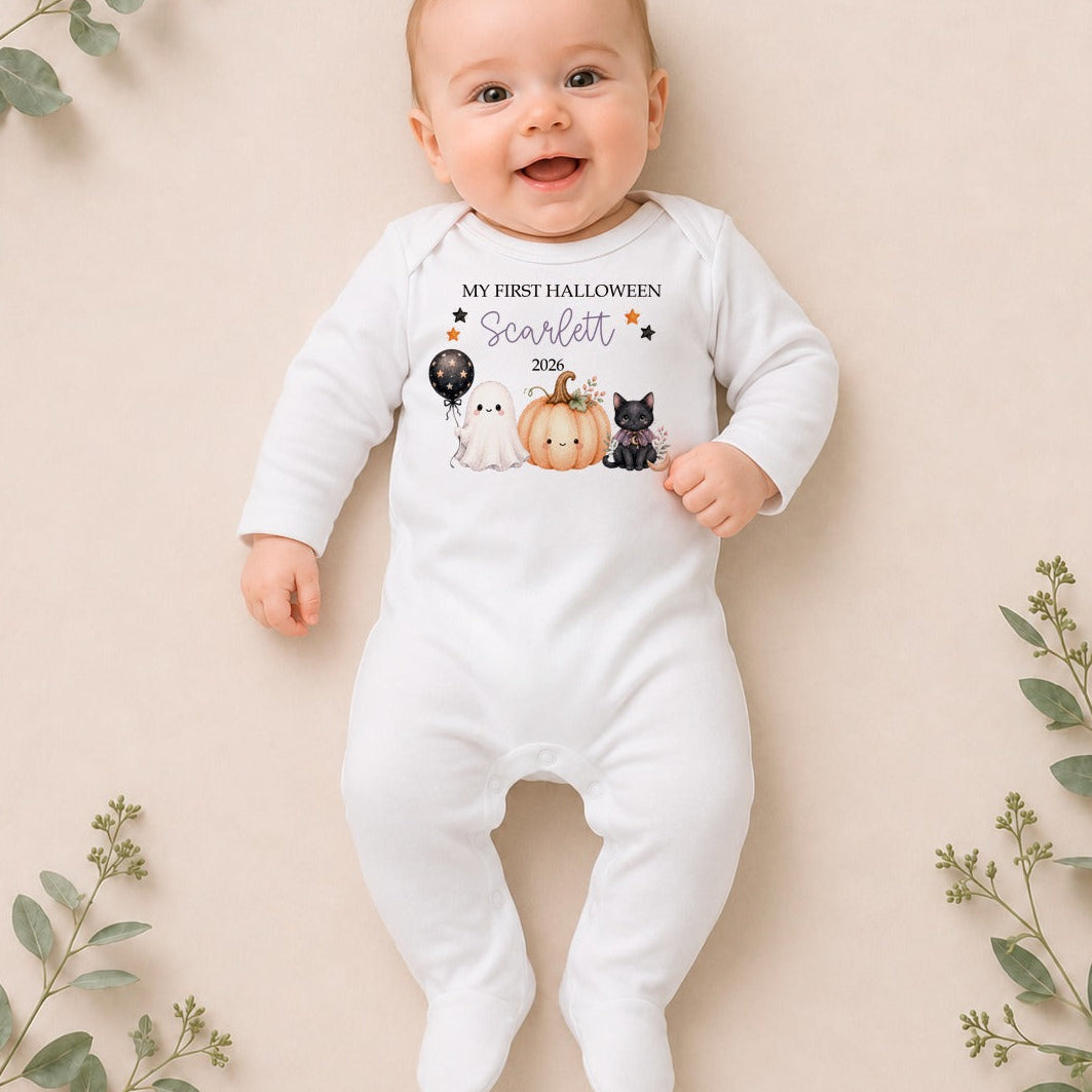 A baby lying on their back wearing a white footed onesie with a graphic that reads 'my first HALLOWEEN scarlet' above a cute illustration of a pumpkin, a black cat, a white cat, and a black dog. The photo is styled with eucalyptus branches on a beige background.