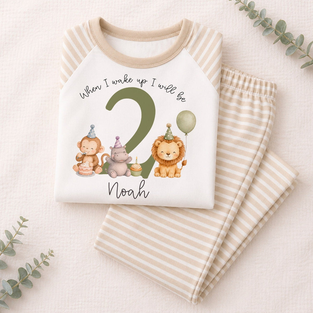 A folded children's pajama set on a light beige textured background with eucalyptus sprigs. The top is cream with beige raglan sleeves and a colorful birthday graphic that reads 'When I wake up I will be 2 Noah' with cartoon animals.