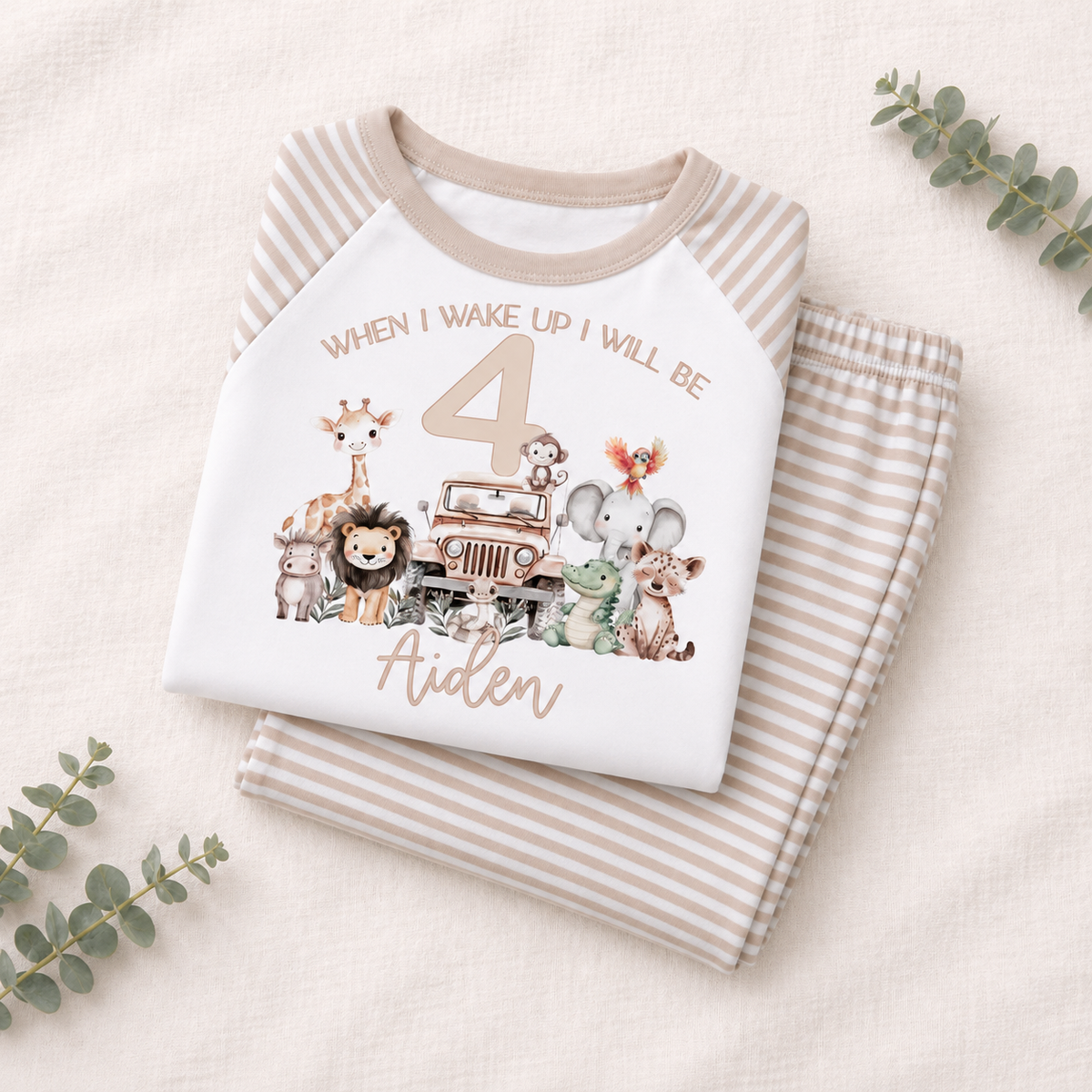 A folded children's pajama set on a light beige fabric background with eucalyptus leaves. The top is a white raglan shirt with beige stripes and a safari-themed 4th birthday graphic reading 'When I Wake Up I Will Be 4 Aiden' with animals and a jeep.