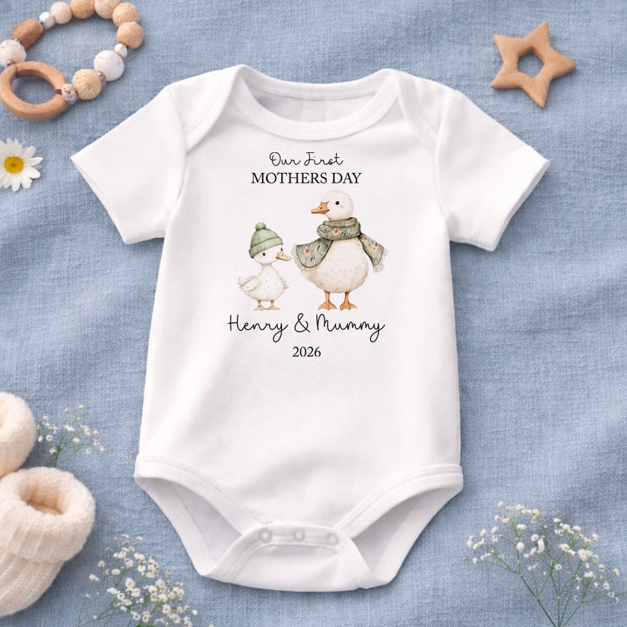 Baby onesie with 'Our First Mothers Day' design on a blue fabric background with baby items.