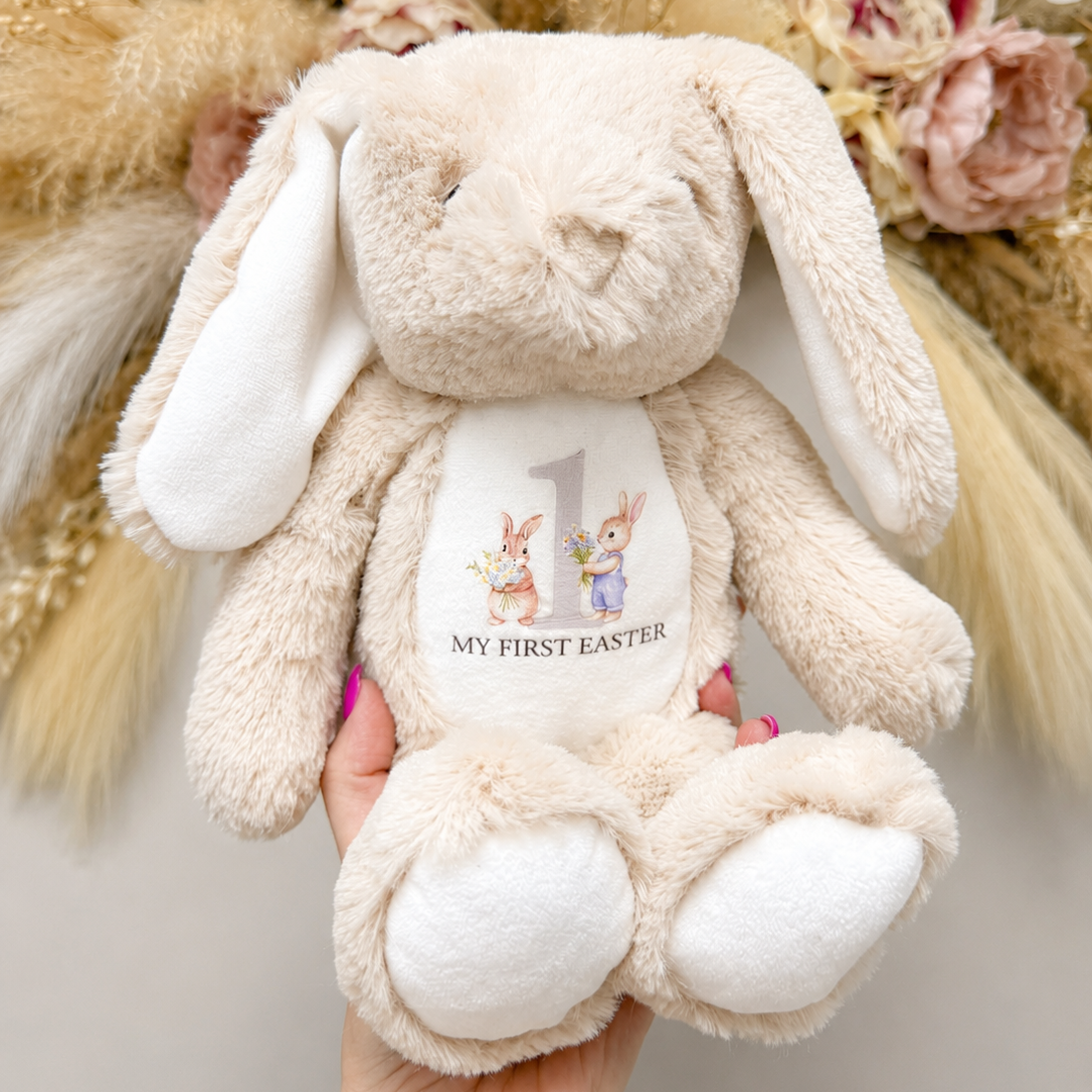 Beige plush bunny with 'My First Easter' label held in front of decorative flowers and plants.