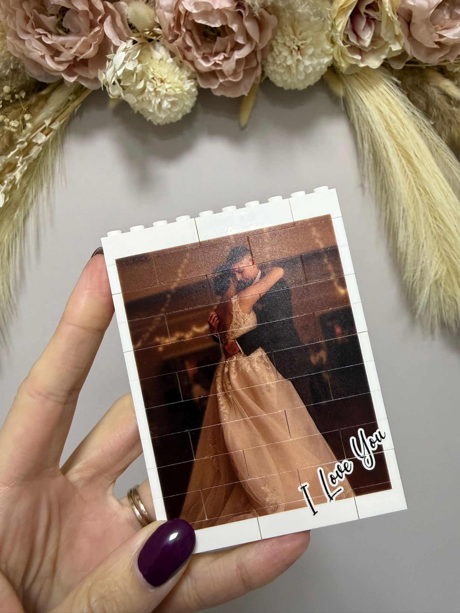 Hand holding a photo of a couple in a dress with 'I Love You' text, surrounded by flowers.