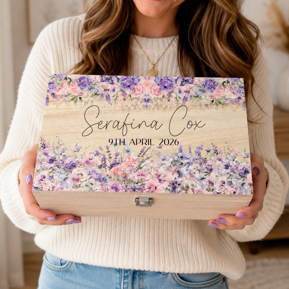 A woman in a cream ribbed sweater holds a personalized wooden keepsake box with a floral watercolor design in purple, pink, and green. The box is customized with the name 'Serafina Cox' and the date '29th April 2026'.