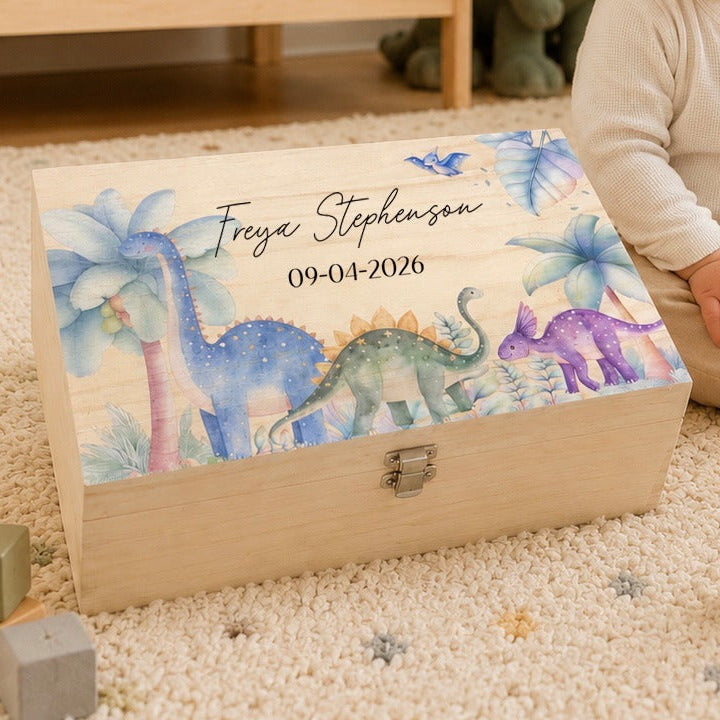 A personalized wooden keepsake box with a colorful dinosaur illustration and the name 'Freya Stephenson' on the lid, placed on a carpeted floor next to a smiling child in a nursery setting.