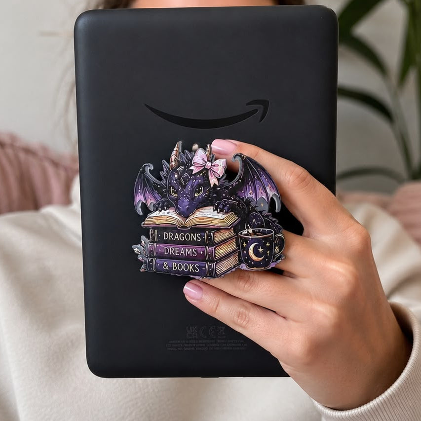 A person holding a black Kindle e-reader in front of their face, featuring a decorative pop-soc style grip with a purple dragon, books, and a camera lens. The person is wearing a cream sweatshirt and sitting on a pink couch with a green plant in the background.