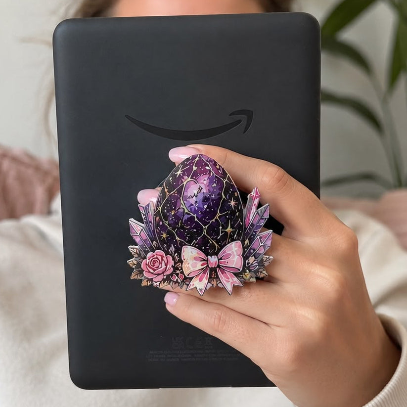 A person in a white sweatshirt holds a black Amazon Kindle e-reader in front of their face, decorated with a purple and pink floral egg-shaped stand featuring a pink bow.