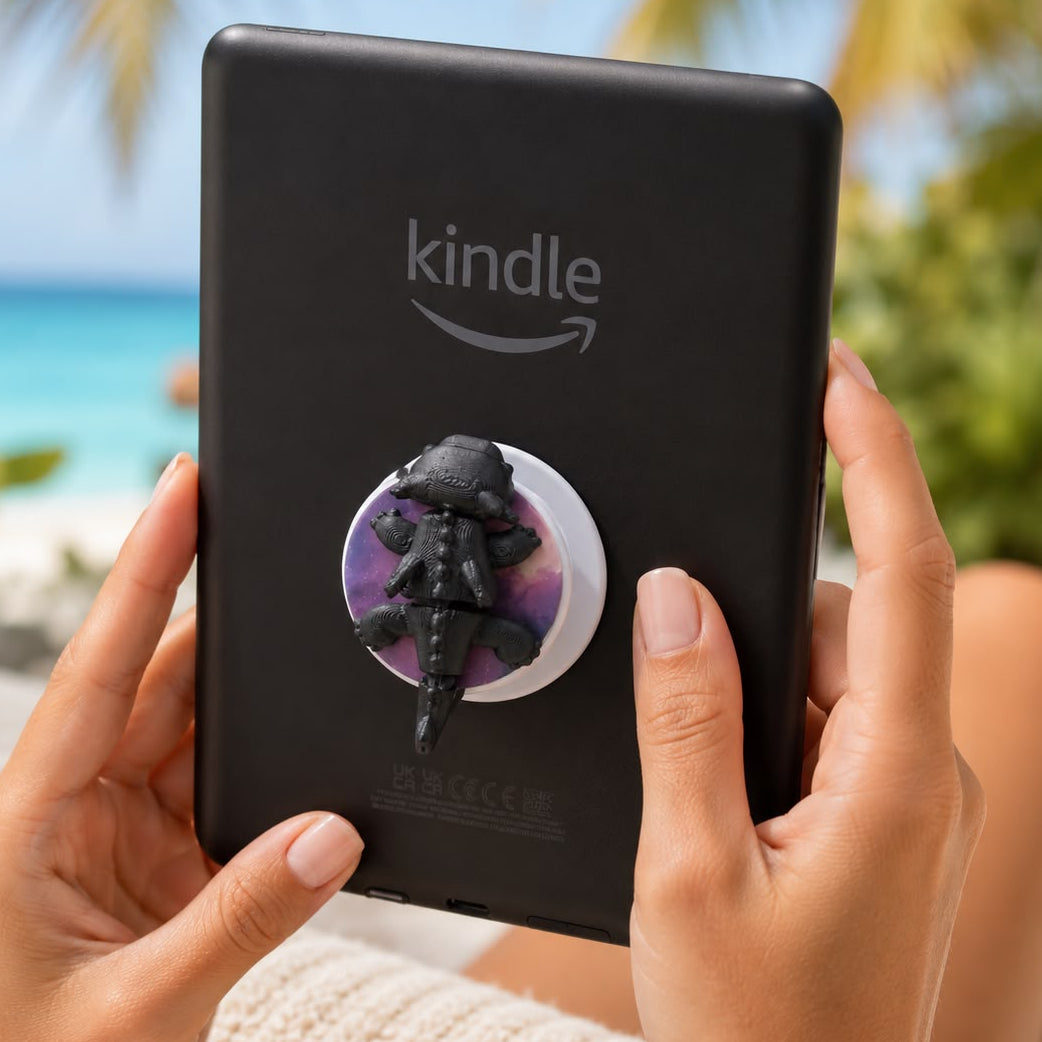 A person holds a black Kindle tablet with a purple and black alligator pop socket against a blurred beach background with palm trees and turquoise water.