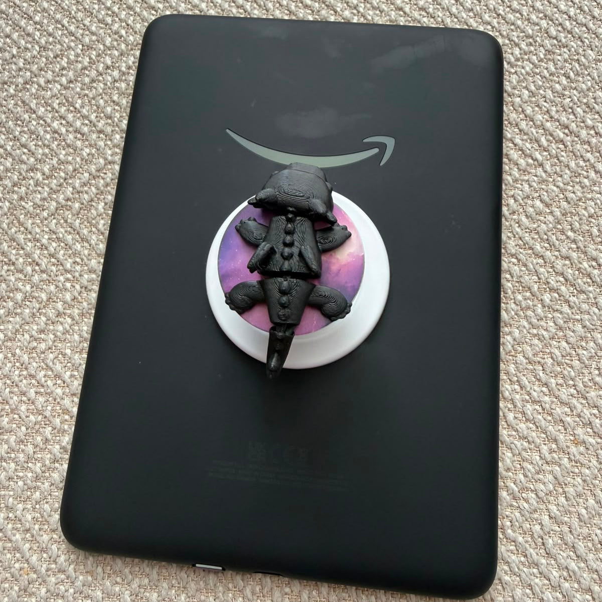 A black Amazon Kindle e-reader device lying on a textured beige carpet, with a small black alligator figurine placed on a round white and purple stand in front of it.