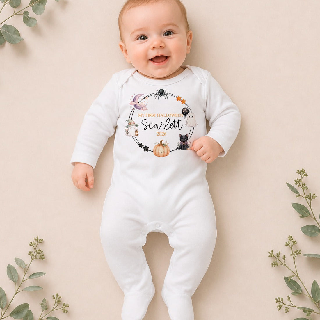 A baby lying on a beige background with decorative eucalyptus branches, wearing a white long-sleeve footed onesie with a Halloween-themed graphic that reads 'My First Halloween Scarlett' with a spider, cat, pumpkin, and hot air balloon design.