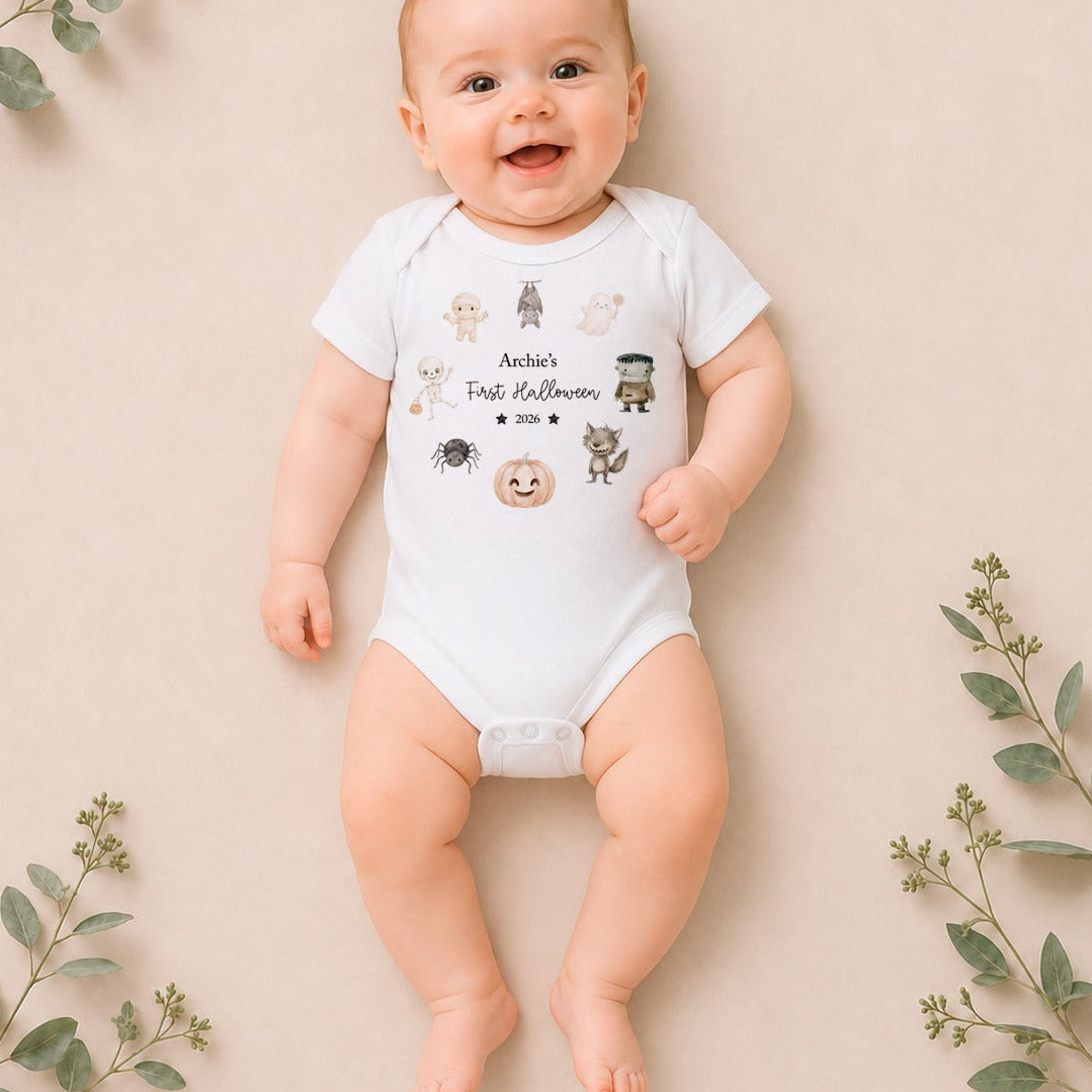 A smiling baby lying on a beige background with eucalyptus leaves, wearing a white short-sleeve bodysuit printed with cute Halloween characters and the text 'Annie's Fake Halloween'.