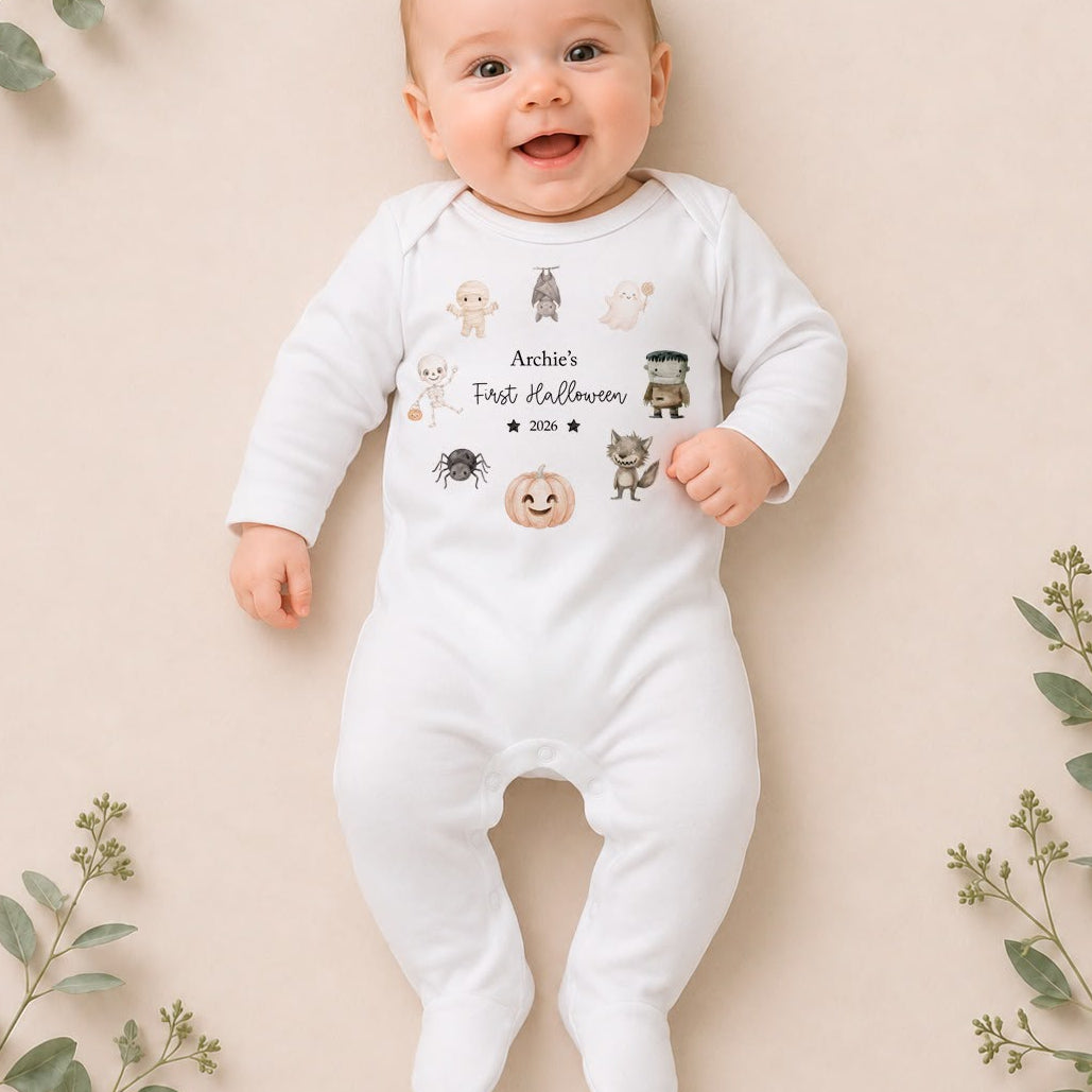 A baby lying on a beige background with decorative eucalyptus branches, wearing a white long-sleeve footed onesie with a 'Annie's First Halloween' graphic print on the chest.