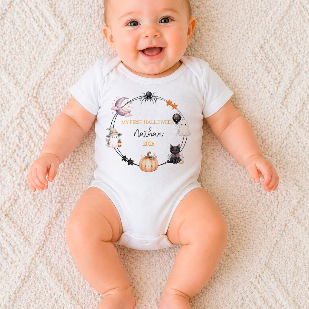 A smiling baby lying on a textured cream blanket, wearing a white short-sleeve bodysuit with a personalized Halloween graphic that reads 'My First Halloween Nathan 2021' with cute spider, cat, and pumpkin illustrations.