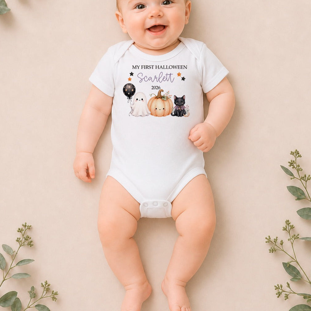 A baby lying on a beige background with decorative eucalyptus branches, wearing a white short-sleeve bodysuit with a Halloween-themed graphic that reads 'My First Halloween: Scarlett 2025' with a pumpkin, black cat, white cat, and black dog.