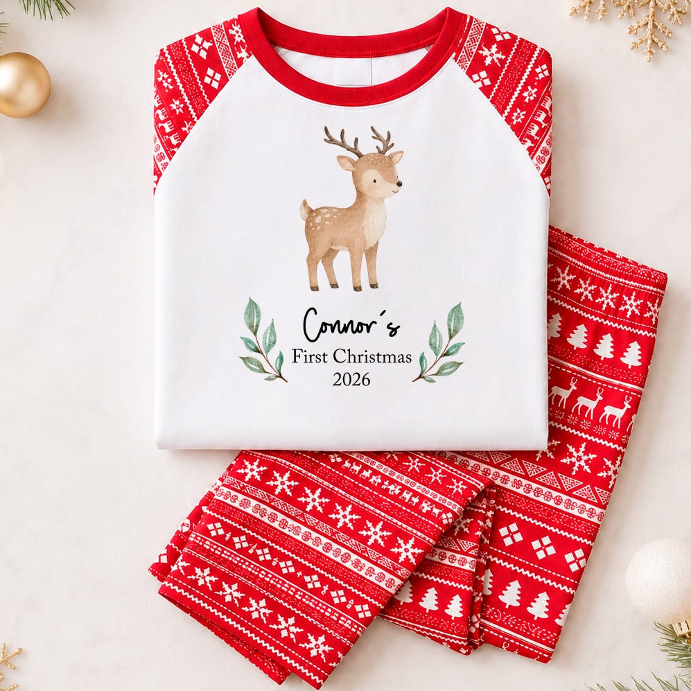 A flat-lay of a two-piece children's pajama set on a light cream background with holiday decorations. The pajama top is white with red raglan sleeves and collar, featuring a printed brown deer and the text 'Connor's First Christmas 2026' with green leaf accents.