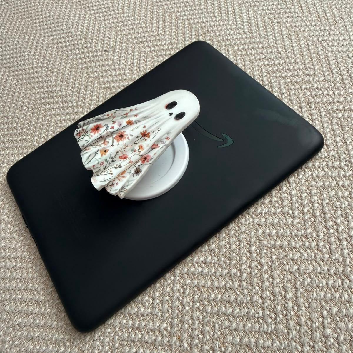 A black tablet case with a white circular magnetic stand holding a white and orange patterned shuttlecock, placed on a textured beige woven surface.