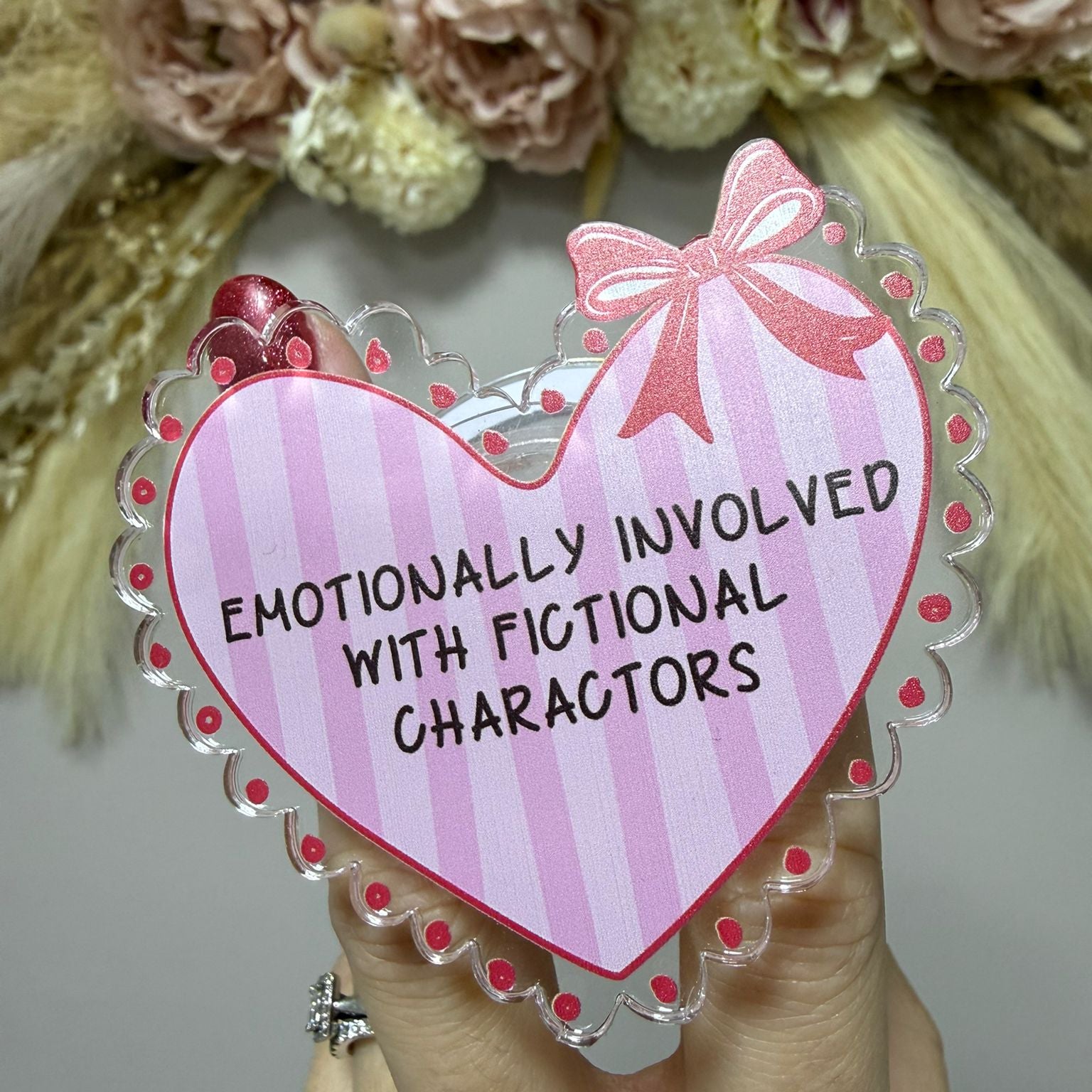 Heart-shaped sign with text 'Emotionally involved with fictional characters' held by a hand, with flowers in the background.