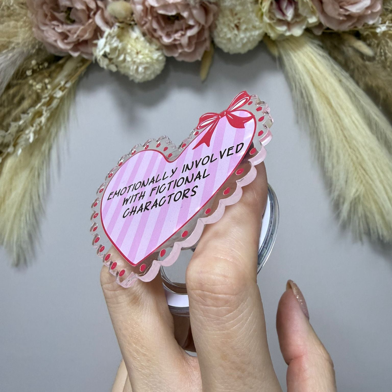 Heart-shaped sticker with text held by a hand in front of a floral wreath.