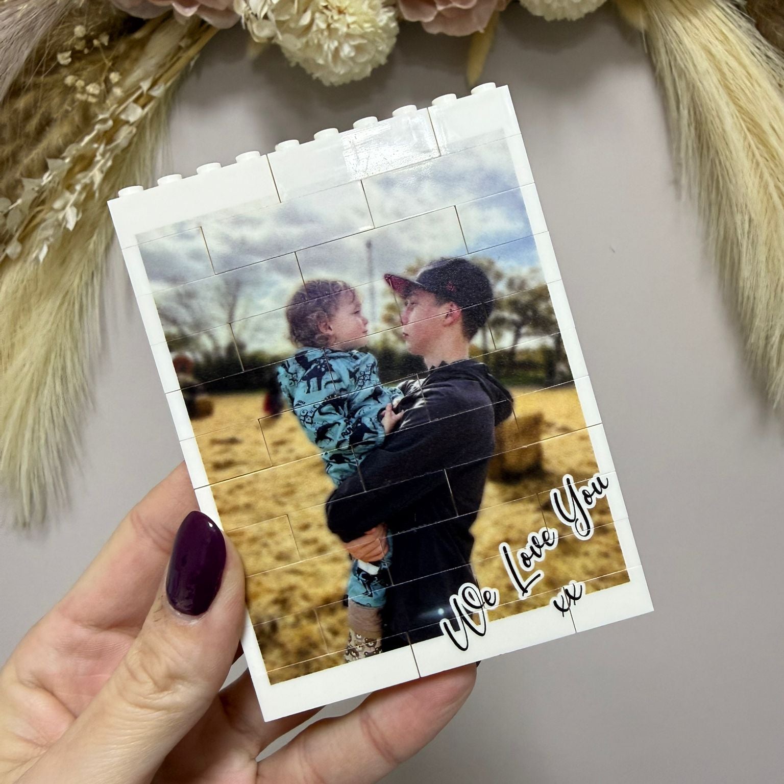 Hand holding a photo card with a printed image of a man and child, text 'We Love You', and decorative flowers in the background.