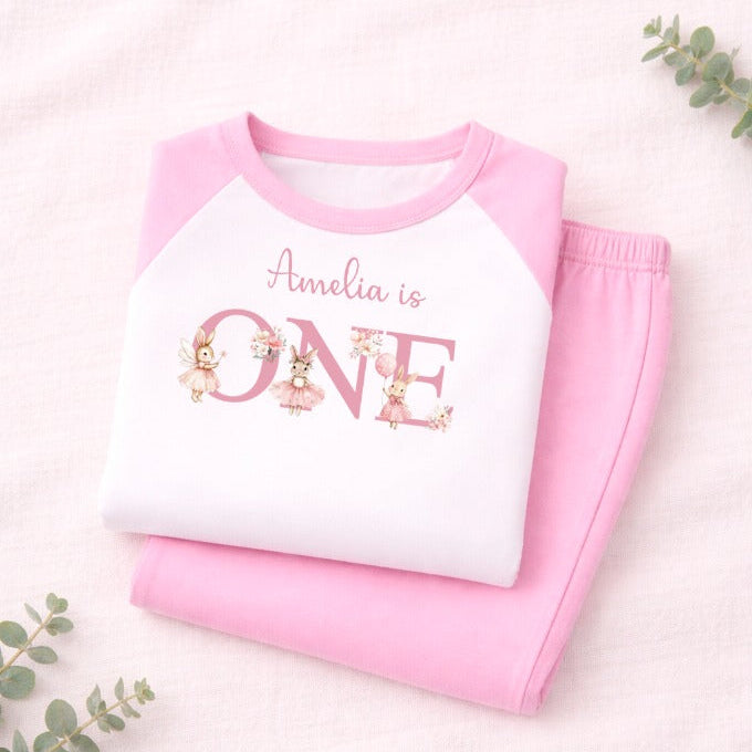 Pink and white children's outfit with 'Amelia is ONE' text on a light background