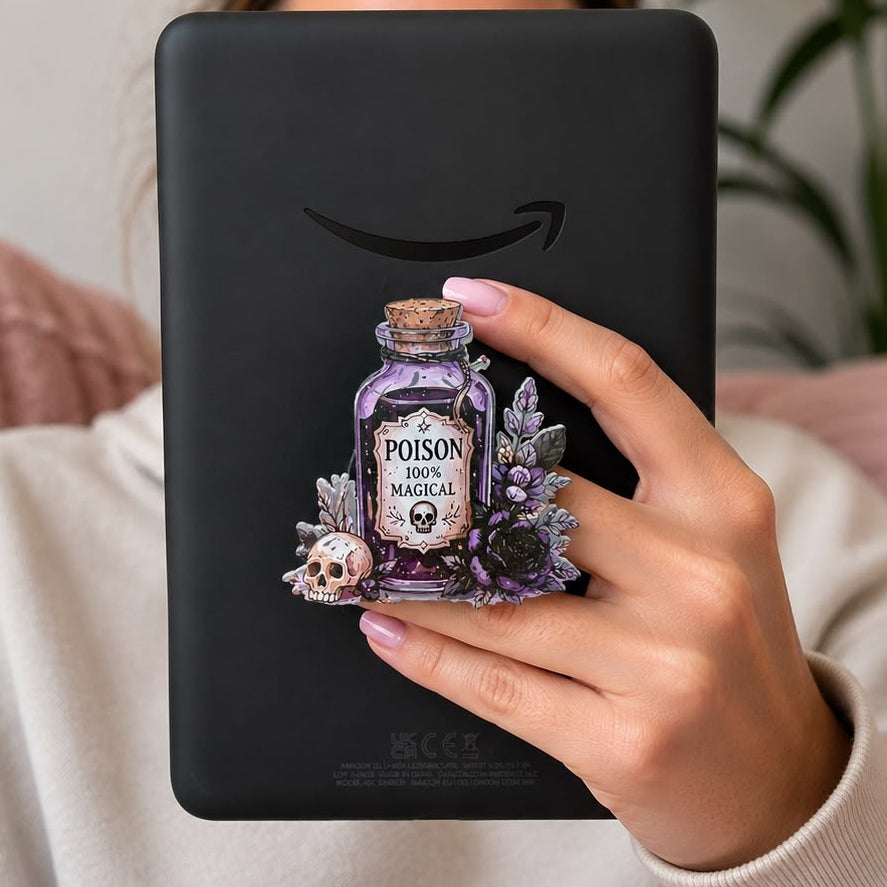 A person holding a black tablet with a decorative purple poison bottle sticker on the back, set against a cozy indoor background with a pink blanket and a green plant.