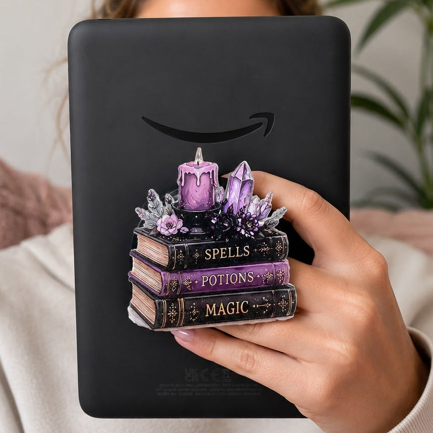 A person in a white sweatshirt holds a black tablet in front of their face, which is obscured by a decorative graphic of stacked books labeled 'Spells', 'Potions', and 'Magic' with purple crystals and a candle. The Amazon smile logo is printed above the graphic.